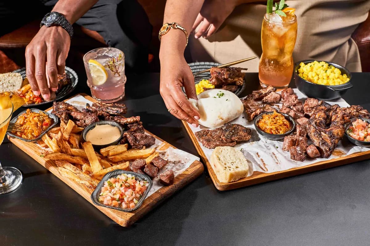 Assorted grilled dishes and cocktails on a restaurant table with a guest reaching for food.