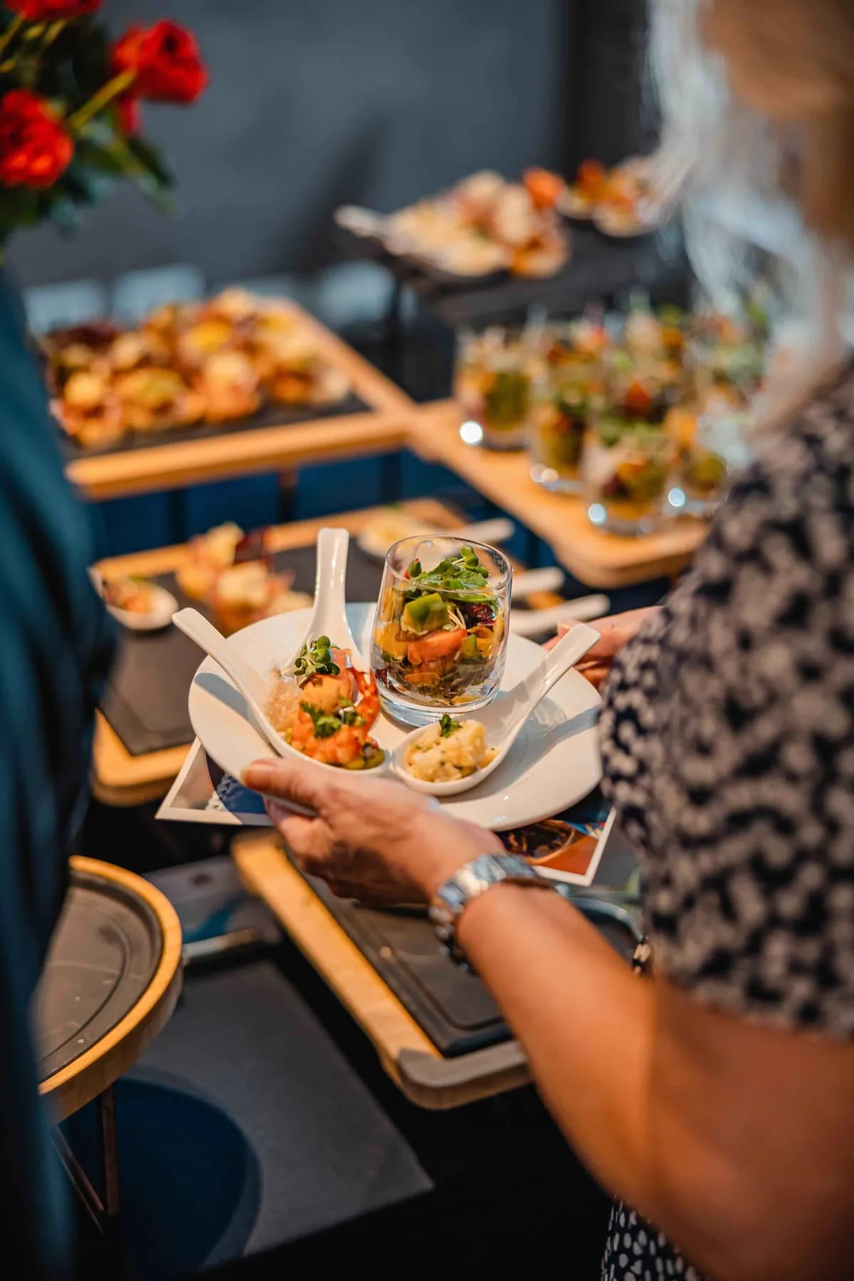 Guest holding a plate of colourful canapés and small dishes at a catered food event, with a buffet spread in the background.
