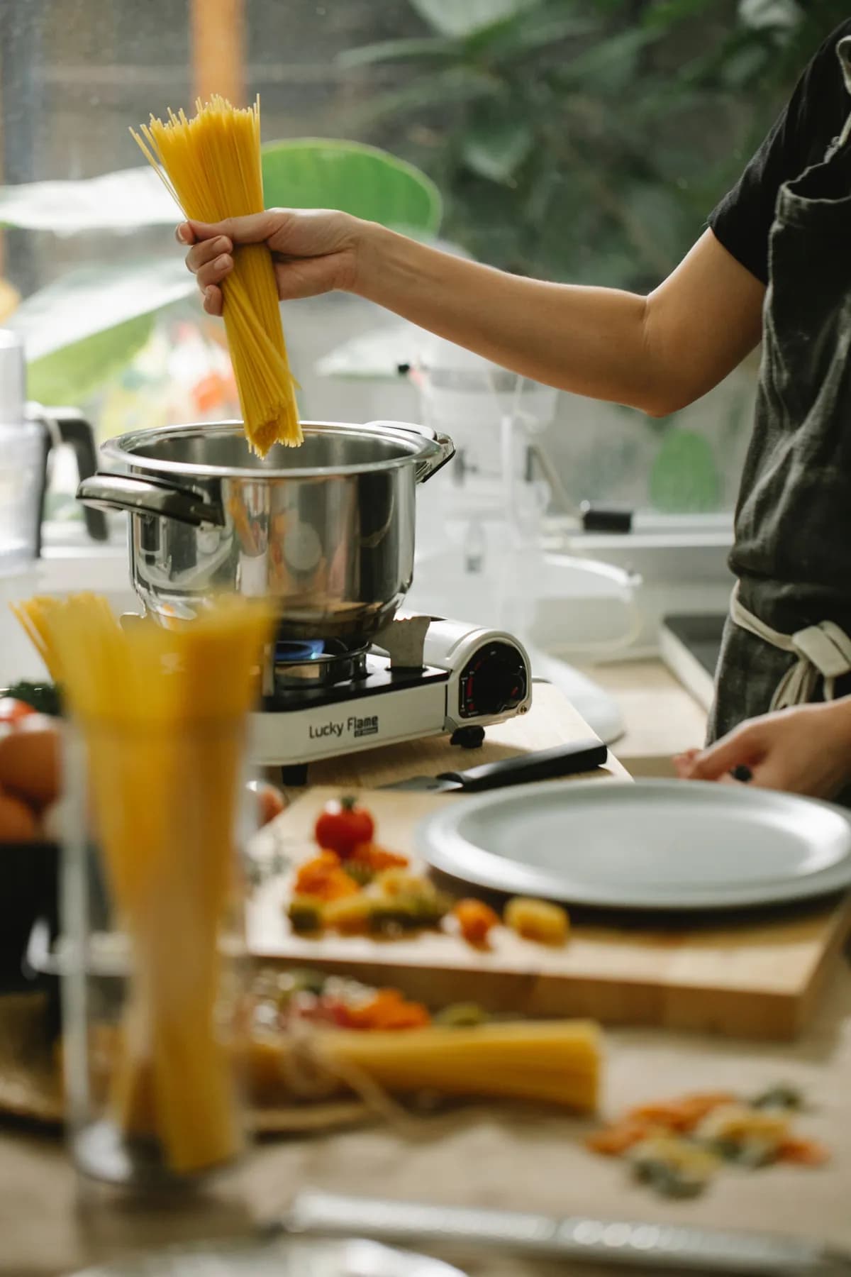 Person adding uncooked spaghetti to a pot of boiling water on a portable gas stove, with pasta ingredients and cooking utensils arranged on a kitchen worktop.
